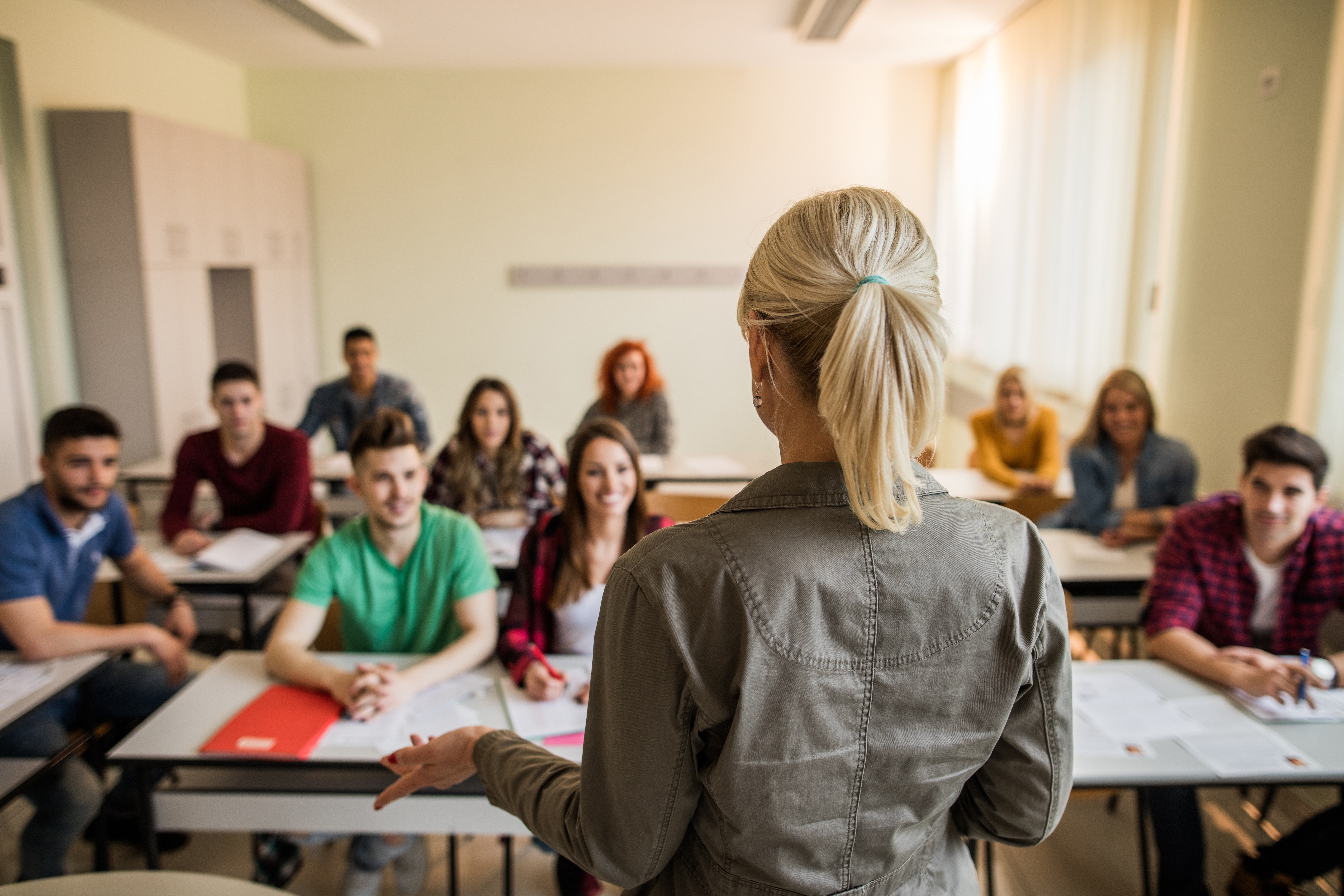 Kvinnelig foreleser står med ryggen til kamera og vendt mot en klasse med rundt ti voksne elever i et klasserom