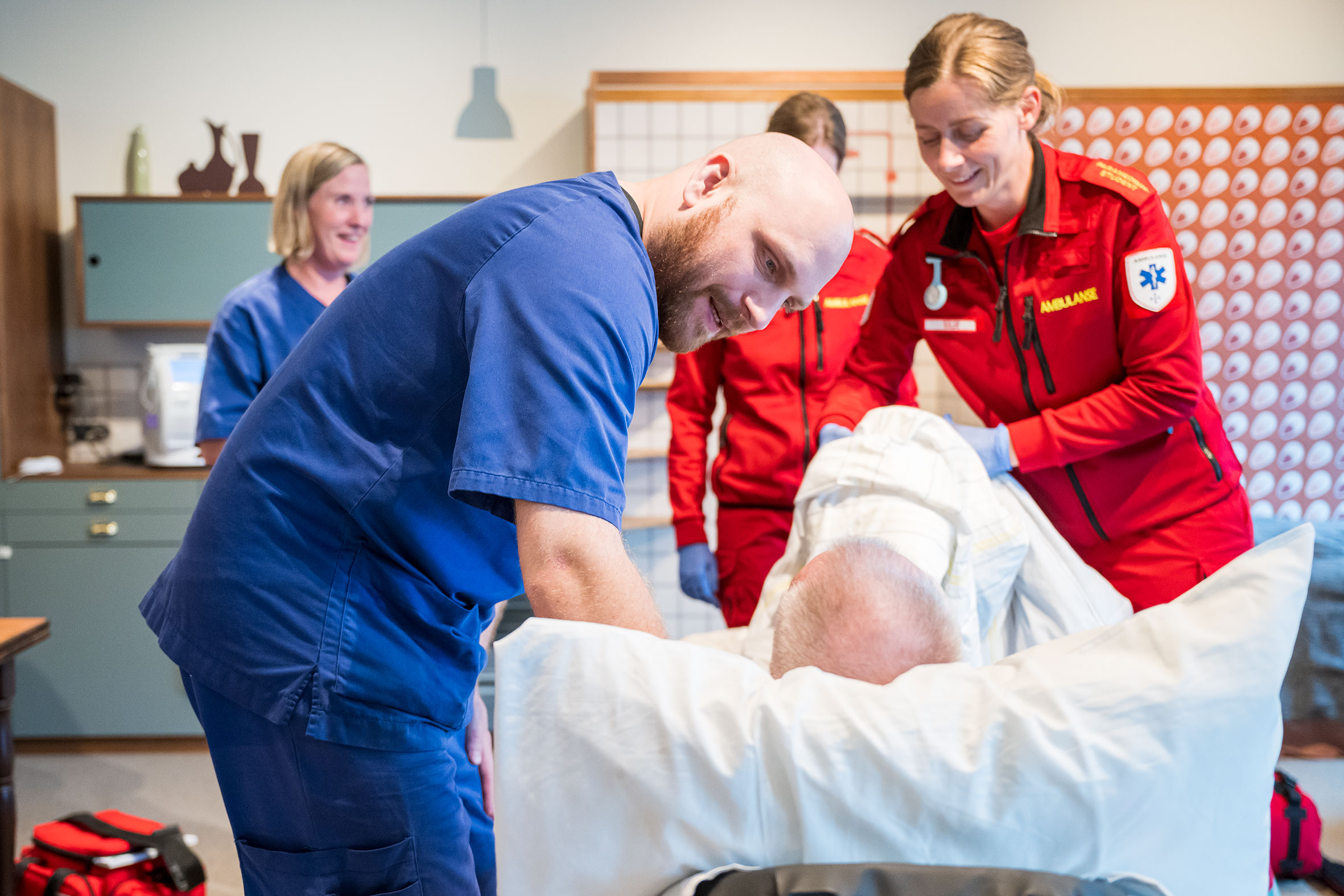 Mannlig sykepleier i blå uniform bøyer seg over pasient sett bakfra i en seng, mens kvinnelig paramedisiner i rød uniform hjelper til.