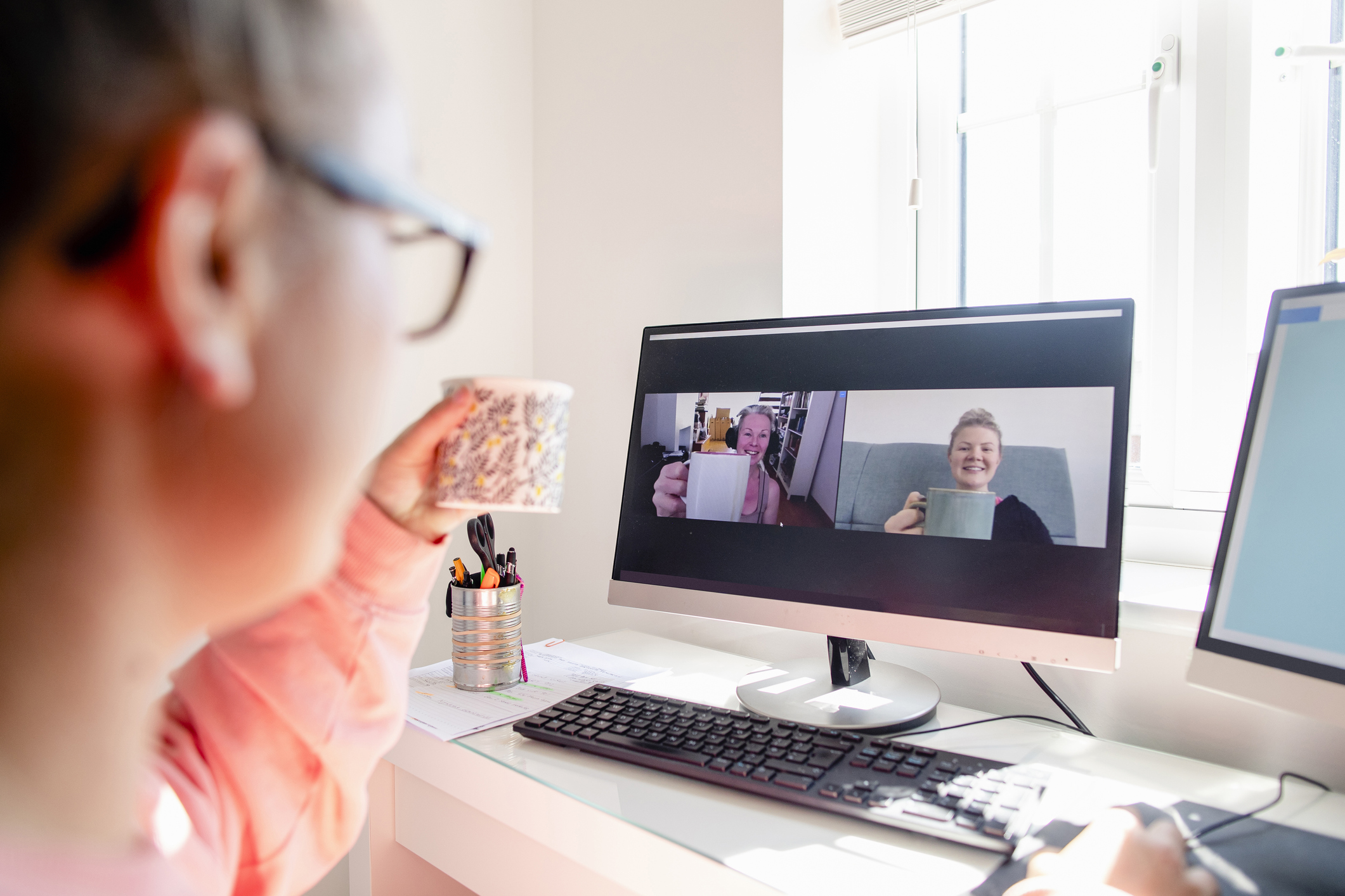 Kvinne skimtes til venstre i bildet. Hun har en kaffekopp i hånda og sitter ved et skrivebord der det er en pc-skjerm med to personer som holder hver sin kaffekopp opp mot henne. Kontorrekvisita på skrivebordet. 