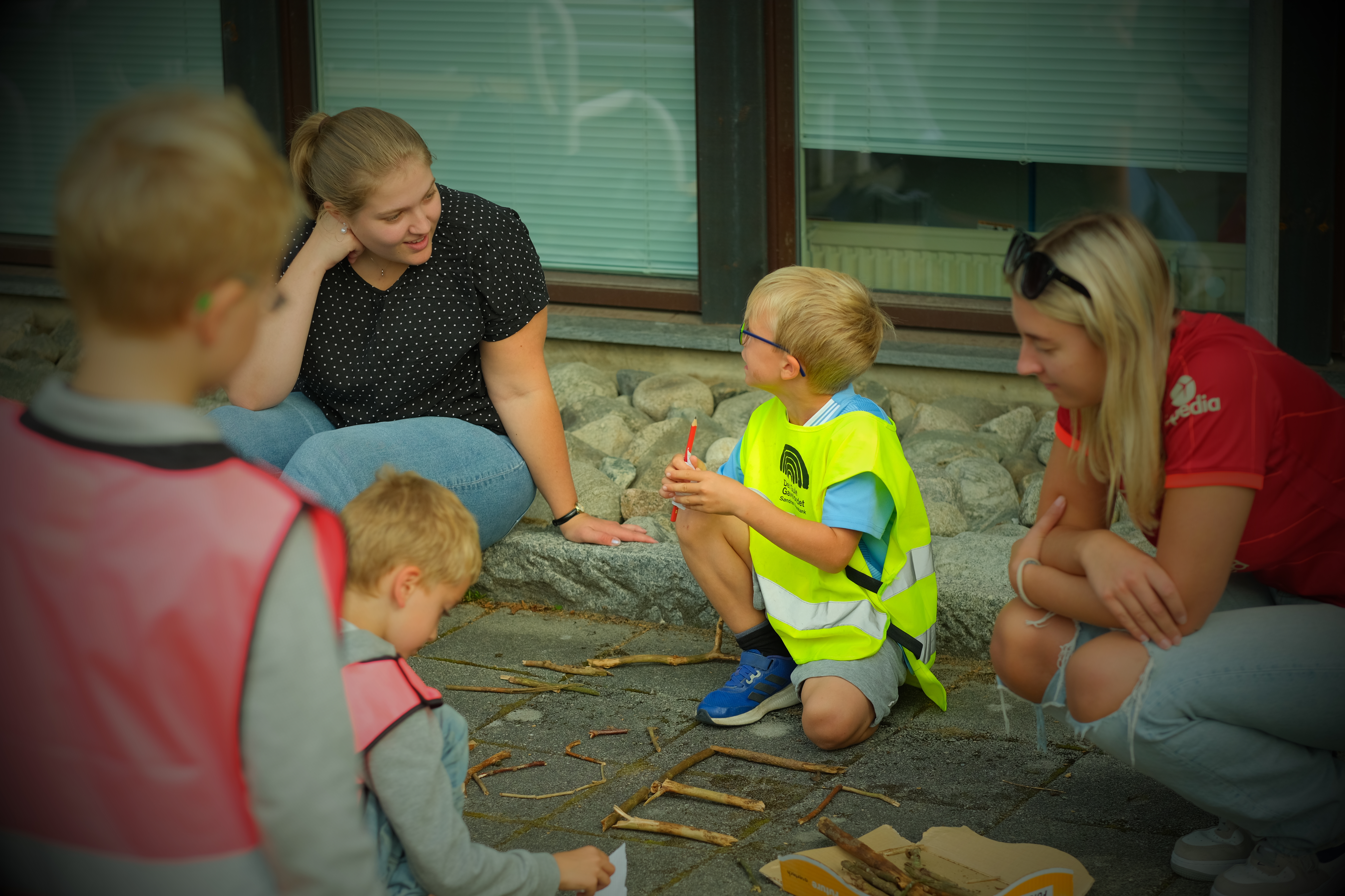 en ung kvinne og en gutt i samtale. De er ute, foran gutten ligger det pinner