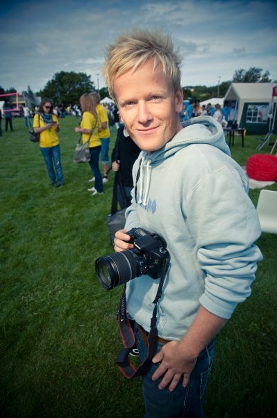 Fotografi av blond mann i lys hettegenser med et kamera i hendene. I bakgrunnen ses mennesker på en gressplen.