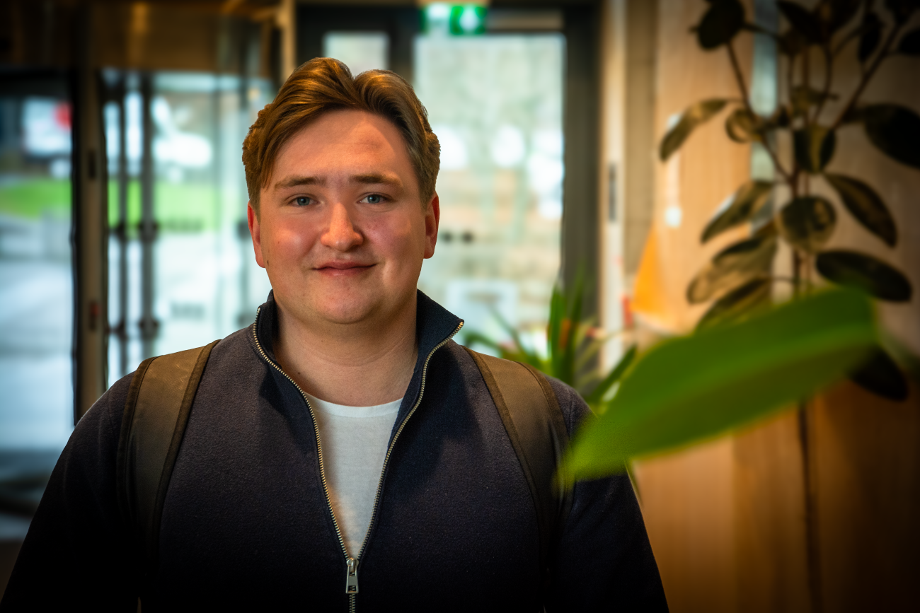 Ung mann med hvit t-skjorte, mørebkå genser med glidelås og sekk på ryggen står inn i en bygning med vinder og svingdører bak seg. Han ser inn i kamera og smiler.