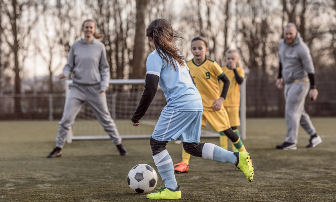 Jenter spiller fotball utendørs. Trener skimtes i bakgrunnen.