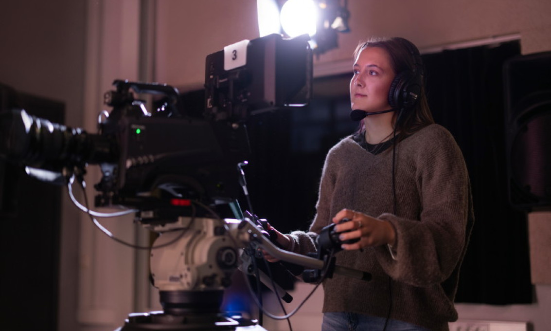 Ung kvinne står bak et stort TV-kamera og styrer det med en spake. Hun har øretelefoner på seg og kikker inn i kameraet.