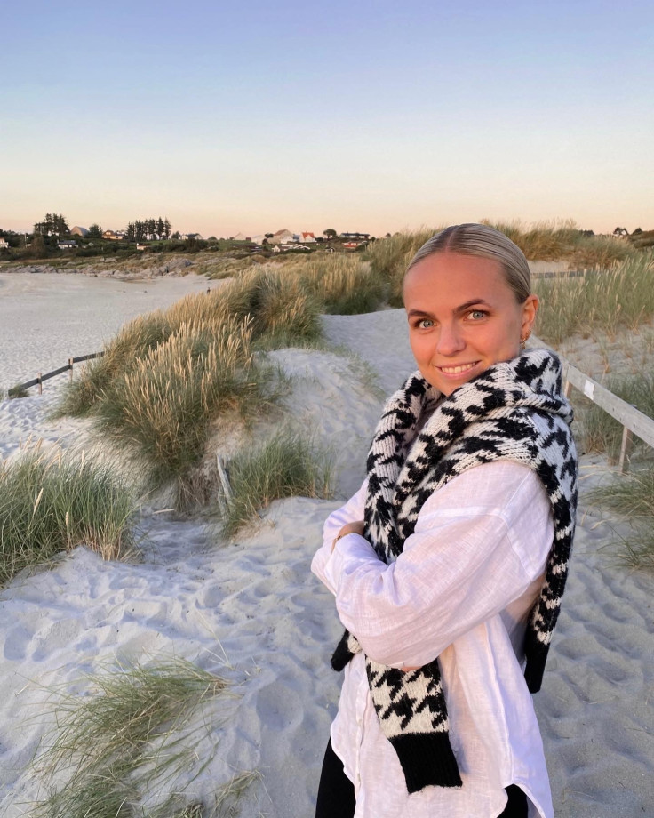ung kvinne med lyst hår står på en sandstrand i solnedgang. Hun har på seg en hvit skjorte og en genser over skuldrene og smiler til kamera.