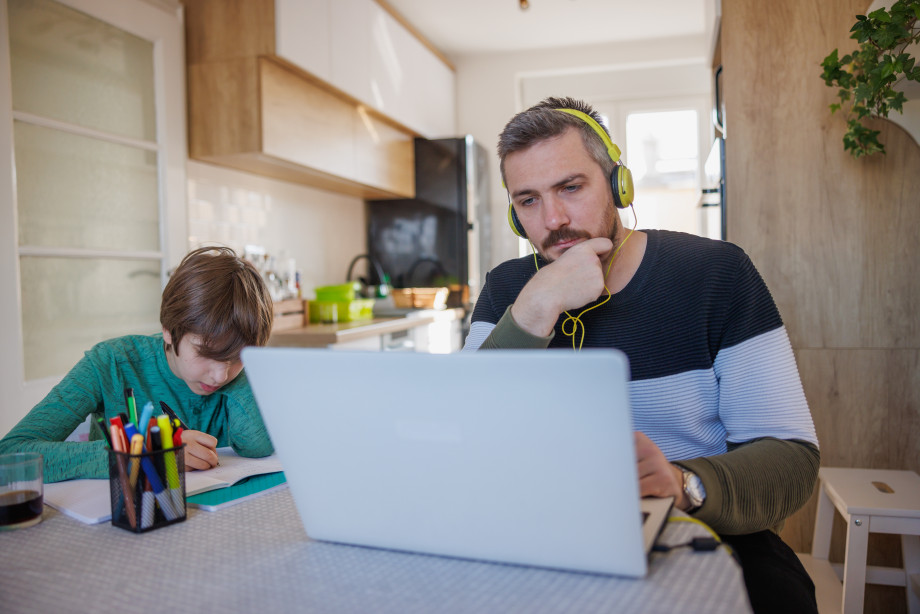 Mann sitter med øretelefoner og datamaskin. Ved siden av sitter et barn og tegner.