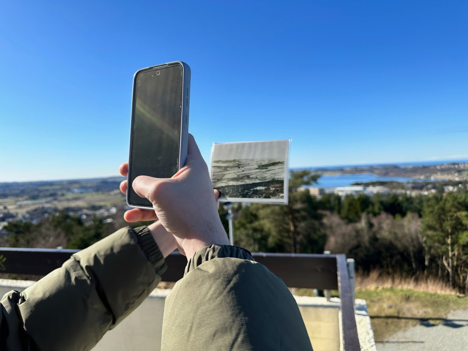 A student at UiS uses a mobile phone and an old photograph to rephotograph a landscape of trees and a lake