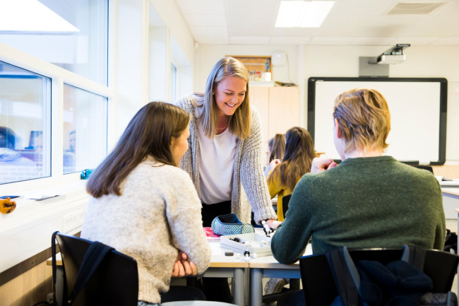 Ung kvinnelige lærerstudent er i et klasserom og hjelper noen elever. Vi ser elevene bakfra.