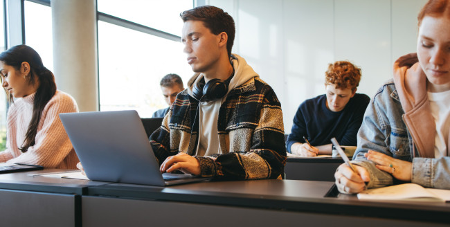 I et klasserom sitter studenter ved pulter i to rader mens de skrive på papir/bærbar datamaskin